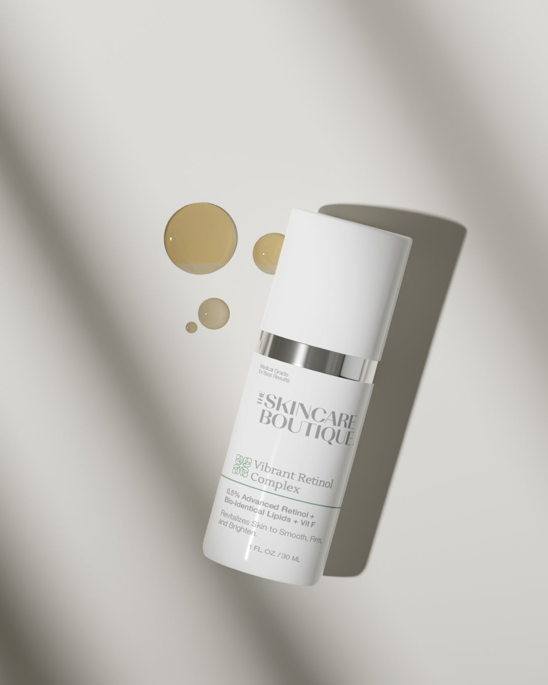 A white skincare bottle labeled 'The Skincare Boutique Vibrant Retinol Complex' with a silver band, photographed on a light gray background with decorative gold droplets and soft shadows.