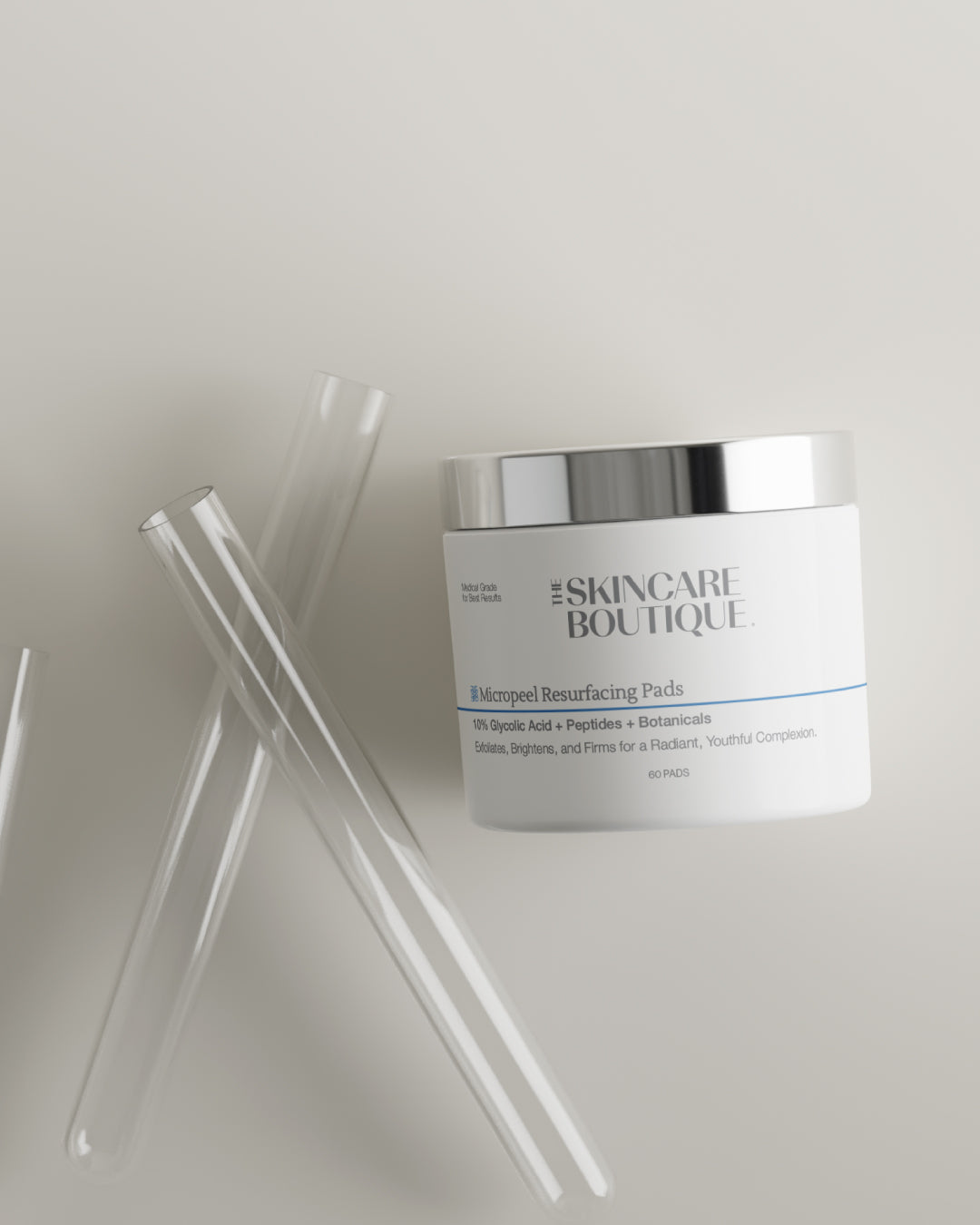 A white cosmetic jar labeled 'The Skincare Boutique' with a reflective silver lid, photographed on a light warm-gray background with three clear glass pipettes placed beside it.