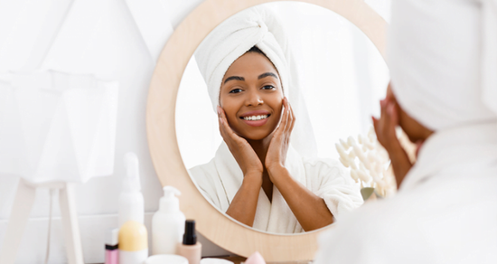 Woman in a white robe with a towel wrapped around her head, looking at herself in a mirror.